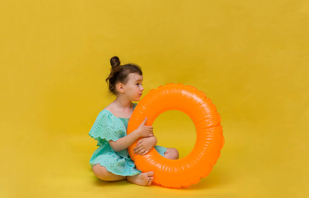 holčička v modrých šatech s oranžovou nafukovací kruh sedí na žlutém pozadí s kopií prostoru - Fotografie, Obrázek
