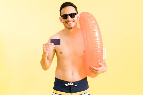 Tohle je nejlepší kreditka. Šťastný pohledný muž s plavkami a slunečními brýlemi šťastný, že platíte kreditní kartou jeho dovolenou na pláži - Fotografie, Obrázek