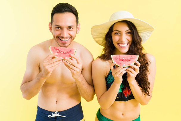 Disfrutando de fruta fresca. Hombre joven latino y mujer caucásica en traje de baño comiendo sandía junto a la piscina durante el verano - Foto, imagen