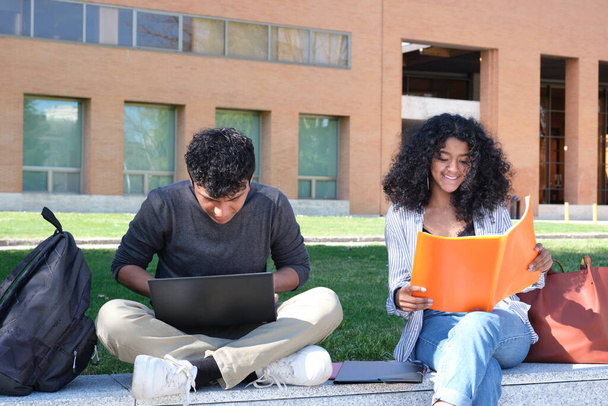 Deux étudiants latins étudiant à partir de leurs notes de cours assis sur un mur à l'extérieur. Vie universitaire sur le campus. - Photo, image