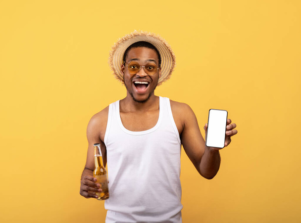 Funky black guy in summer clothes holding cellphone with blank screen and bottle of beer on yellow background, mockup - Foto, imagen