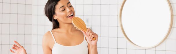 mujer afroamericana joven con los ojos cerrados cantando con cepillo de pelo en el baño, pancarta - Foto, Imagen