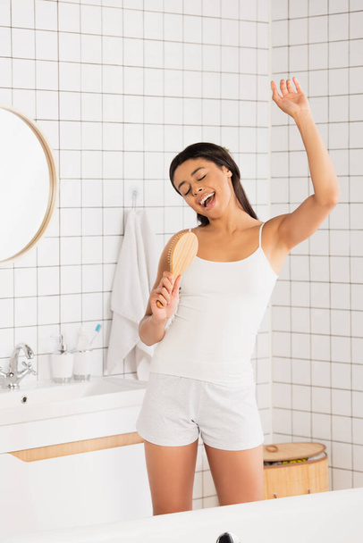 jovem afro-americana em roupas brancas cantando com escova de cabelo no banheiro - Foto, Imagem