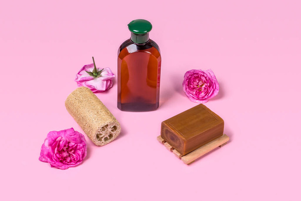 Φυσικό σαπούνι, loofah loofah και ένα μπουκάλι καλλυντικού προϊόντος σε ροζ φόντο με τριαντάφυλλα με χώρο αντιγραφής. Φυσικά βιολογικά καλλυντικά προσώπου και σώματος. Έννοια φροντίδας δέρματος. - Φωτογραφία, εικόνα