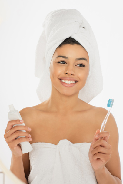 positivo jovem afro-americano mulher envolto em toalhas segurando escova de dentes e pasta de dentes no banheiro - Foto, Imagem