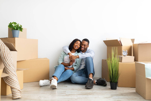 Amare i coniugi neri che si abbracciano tra scatole di carta forniture in movimento con oggetti in appartamento vuoto - Foto, immagini