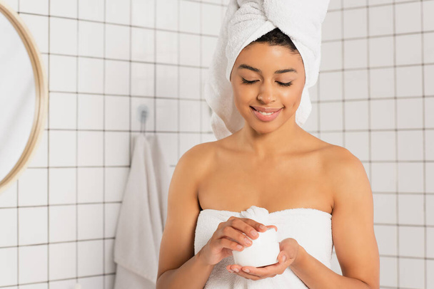 smiling young african american woman holding jar with cream in bathroom - Foto, afbeelding
