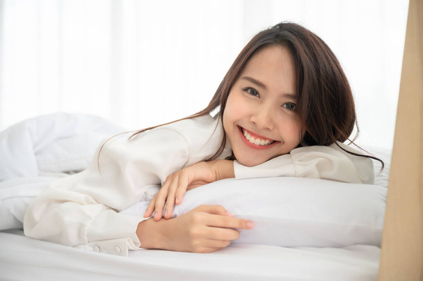 Joven mujer asiática despertando en la mañana en la cama. Ella mostrando la cara hacia fuera la manta y sonreír sintiéndose feliz - Foto, imagen