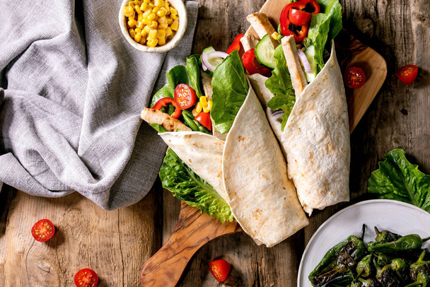 Traditional mexican tortila wrap with pork meat and vegetables on wooden cutting board decorated with white textile napkin corn and grilled jalapenos over brown wooden background. Top view, flat lay - Φωτογραφία, εικόνα