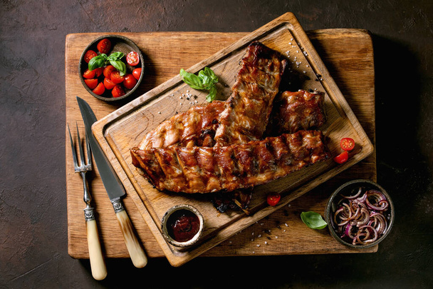 Grilled pork bbq ribs served with marinated onions, cherry tomatoes, basil and barbeque sauce on wooden cutting board over dark brown texture background. Vintage cutlery. Top view, flat lay - Φωτογραφία, εικόνα