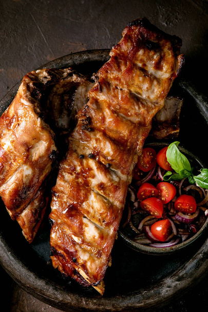 Grilled pork bbq ribs in ceramic plate served with marinated onions, cherry tomatoes, basil over dark brown texture background. Top view, flat lay. Close up - Φωτογραφία, εικόνα