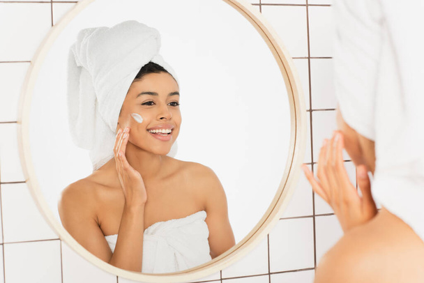 uśmiechnięta młoda Afroamerykanka owinięta w ręczniki nakładająca krem na twarz w pobliżu lustra w łazience - Zdjęcie, obraz