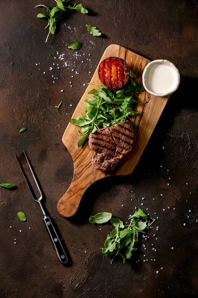 Grilled beef steak served with creamy sauce, grilled vegetables tomato and arugula salad on wooden cutting board with meat fork over dark brown texture background. Top view, flat lay - Φωτογραφία, εικόνα