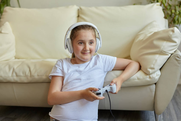 家でソファに座っている間に画面と笑顔を見て、ヘッドフォンでかなり小さな女の子は、ゲームコンソールを果たしている - 写真・画像