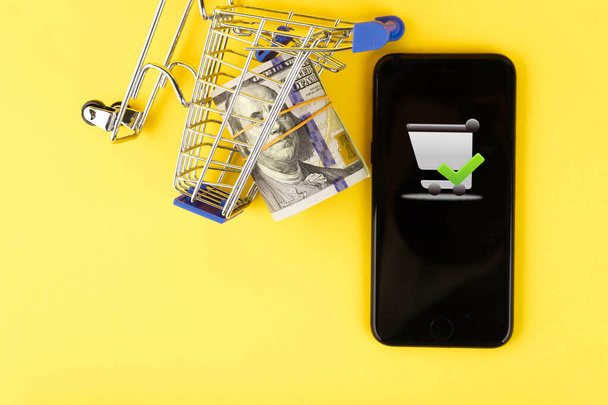 Online vásárlás, telefon és bevásárlókocsi koncepció 100 dolláros bankjegyekkel sárga háttérrel. Fénymásolási hely. - Fotó, kép