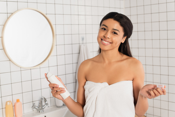 young african american woman wrapped in towel holding deodorant in bathroom - Foto, imagen