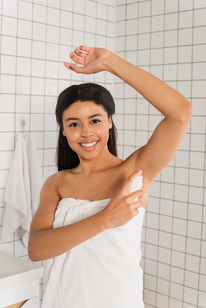 sorridente giovane donna afroamericana avvolta in asciugamano spruzzando con deodorante in bagno - Foto, immagini