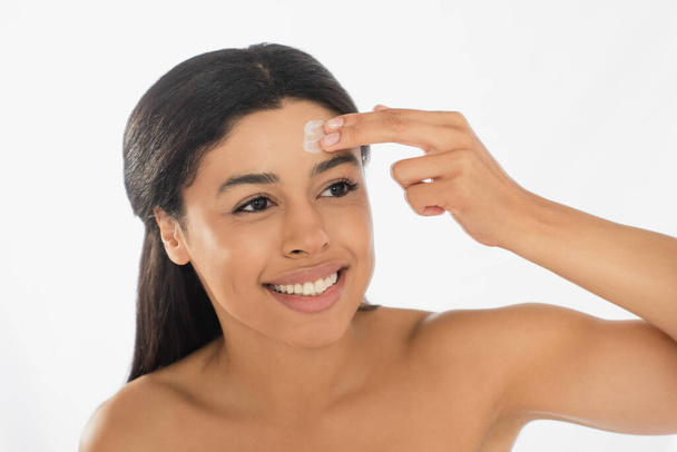 sonriente joven afroamericana mujer aplicando crema en la frente sobre fondo blanco - Foto, Imagen