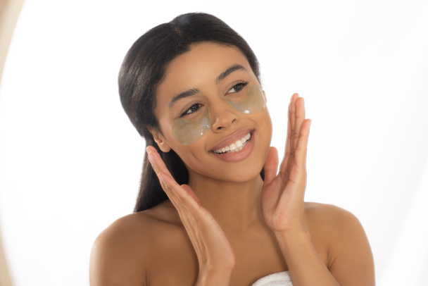 heureuse jeune femme afro-américaine tenant la main près du visage avec des patchs dans la salle de bain - Photo, image