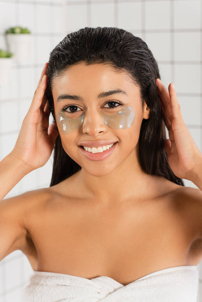 улыбающаяся молодая африканская американка с повязками на глазу, держащая руки за голову в ванной - Фото, изображение