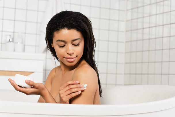 young african american woman sitting in bathtub and applying cream on arm in bathroom - Fotó, kép
