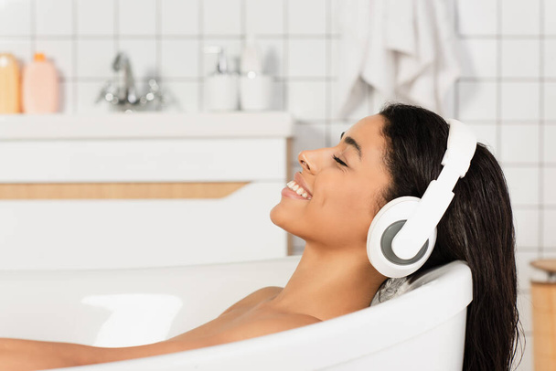 glückliche junge Frau, die mit geschlossenen Augen in der Badewanne liegt und über Kopfhörer Musik hört - Foto, Bild