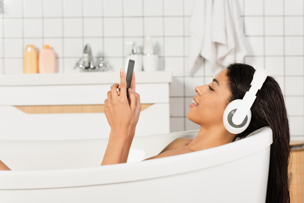 puolella näkymä hymyilevä nuori nainen kuuntelee musiikkia kuulokkeet katsomalla matkapuhelin kylpyhuoneessa - Valokuva, kuva