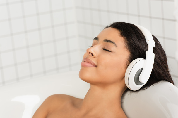 jeune femme souriante avec les yeux fermés se reposant et écoutant de la musique dans les écouteurs dans la salle de bain - Photo, image