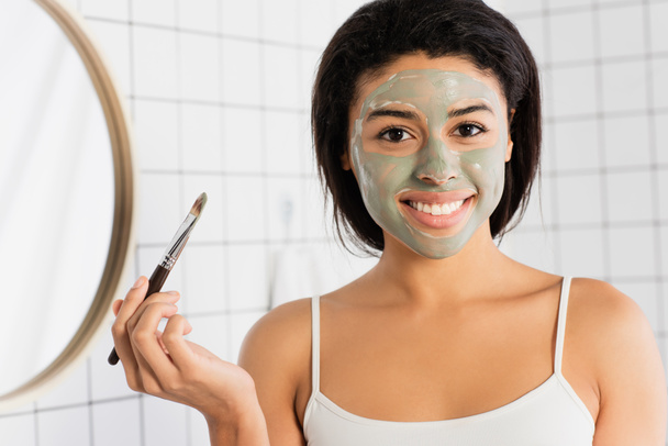 sonriente mujer afroamericana con máscara de barro en la cara con cepillo cosmético y mirando a la cámara en el baño - Foto, Imagen
