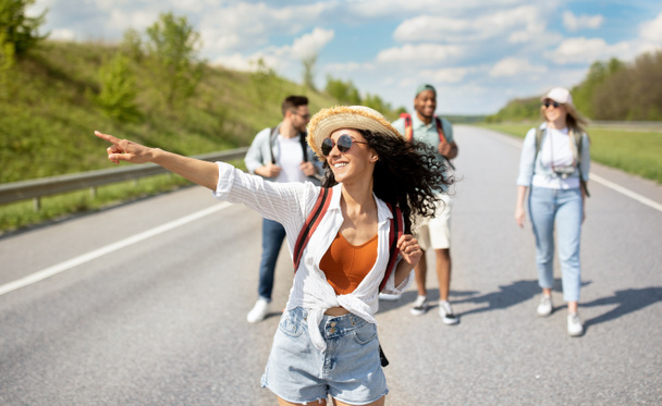 Joven mujer caucásica con sus amigos multirraciales haciendo autostop en la carretera, viajando por autostop, al aire libre - Foto, imagen