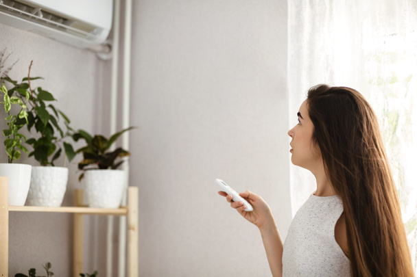 Jeune femme ajuste la température du climatiseur à l'aide de la télécommande dans la pièce avec beaucoup de plantes à la maison - Photo, image