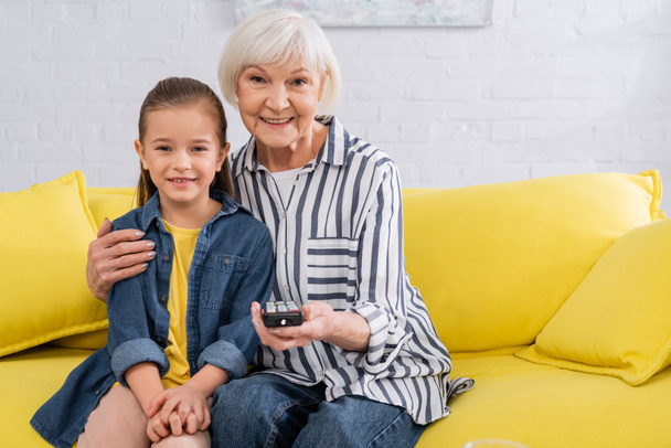 Grandmother with remote controller hugging child on couch  - Foto, Imagem