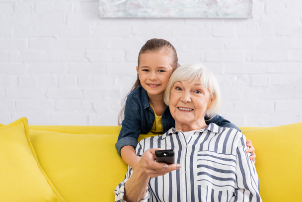 Enkelin umarmt Oma mit Fernbedienung zu Hause  - Foto, Bild