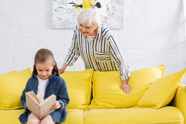 Lächelnde Oma steht neben Kind und liest Buch zu Hause  - Foto, Bild