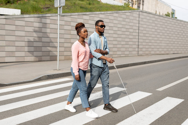 Joven mujer negra asistiendo a un chico millennial con discapacidad visual con bastón cruzando la calle de la ciudad. Concepto de discapacidad visual - Foto, Imagen