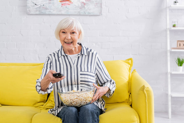 Mujer mayor positiva sosteniendo mando a distancia y palomitas de maíz en casa  - Foto, Imagen