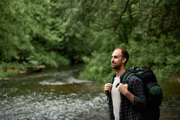 川のほとりに立って自然を楽しむ大きなハイキングバックパックを持つ男のサイドビュー。ハイキング旅行の概念は、荒野の中で野外生活の冒険、休暇をさまよう. - 写真・画像