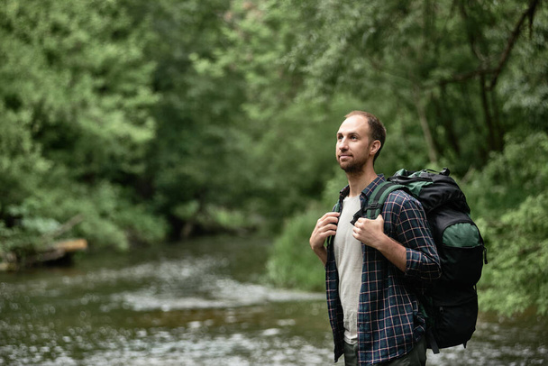 Hiker with a large hiking backpack standing on the bank of the river and enjoying nature. The concept of hiking trips, wanderlust lifestyle vibes of adventure, vacations outdoors in the wilderness. - Photo, Image