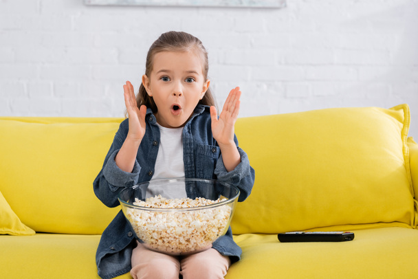 Amazed child looking at camera near popcorn and remote controller  - Photo, image