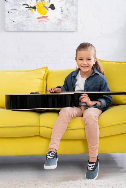 Positive kid smiling at camera near acoustic guitar on couch  - Фото, изображение