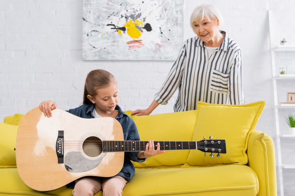 Elderly woman standing near couch and kid holding acoustic guitar  - Foto, Imagem