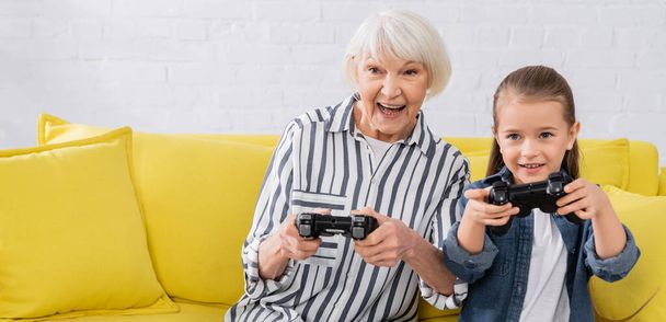KYIV, UKRAINE - JANUARY 11, 2021: Positive granny playing video game near granddaughter with joystick, banner  - Foto, Imagen