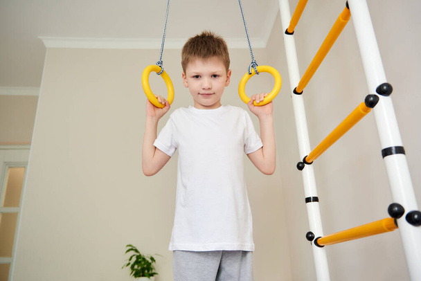 Jongen doet gymnastiek oefeningen indoor. 7 jaar oud kind dat op sportuitrusting speelt. Jongen in sportkleding doet oefeningen op sportringen op een Zweedse muur. Kindersport. - Foto, afbeelding