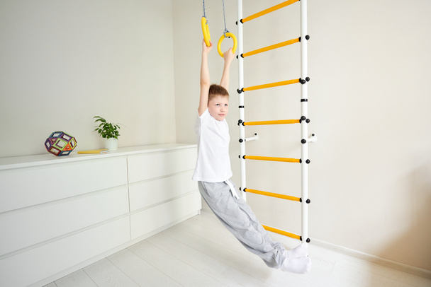 De jongen hangt aan zijn handen en doet fysieke oefeningen om zijn benen op te heffen, waarbij hij de cortex spieren inschakelt. Fysieke, Hand-en Oogcoördinatie, Sensorische, Motor Skills ontwikkeling, Home workout concept - Foto, afbeelding