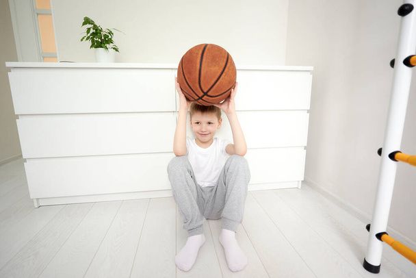 Mały uśmiechnięty uczeń siedzący na podłodze w swoim białym pokoju, trzymający koszykówkę nad głową, patrzący i marzący o zajęciach sportowych. Portret dziecka bawiącego się koszykówką. - Zdjęcie, obraz