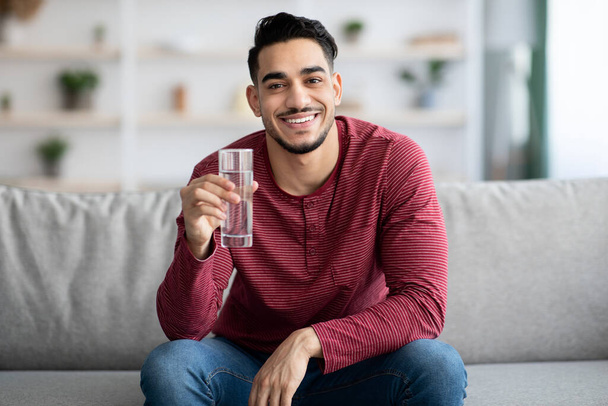Cheerful arab guy sitting on couch, drinking water, copy space - Fotó, kép