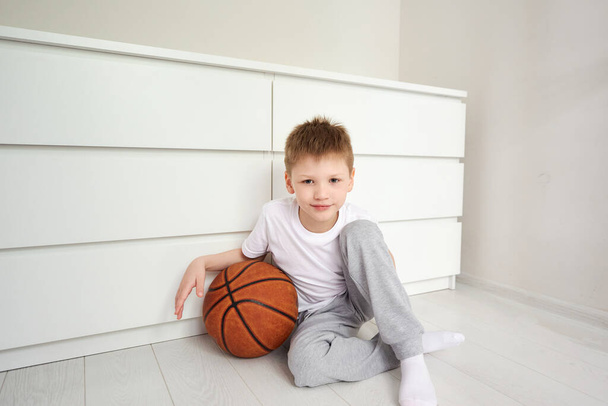 Portret małego uśmiechniętego ucznia patrzącego w kamerę siedzącego na podłodze w swoim białym pokoju, z koszykówką w pobliżu. Sport i zajęcia dla dzieci koncepcja. Kwarantanna w domu. - Zdjęcie, obraz
