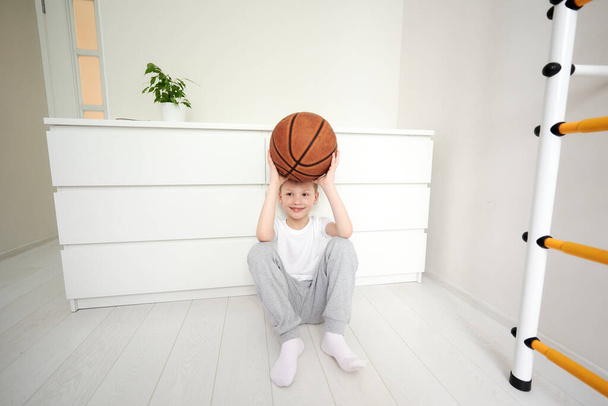 Malý usměvavý školáček sedí na podlaze u svého bílého pokoje, drží nad hlavou basketbal, dívá se a sní o sportovních aktivitách. Karanténa doma. - Fotografie, Obrázek