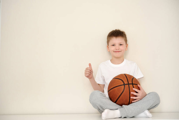 バスケットボールをしている彼の部屋に座っている小さな笑顔の少年のポートレットは、スポーティーなライフスタイルを選択する兆候として親指を上げています。白地だ。テキストのスペース - 写真・画像