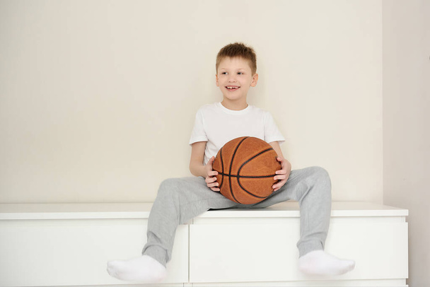 Cute smiling boy in casual clothes sitting on the dresser in his room holding a basketball. White background. Space for text - Φωτογραφία, εικόνα